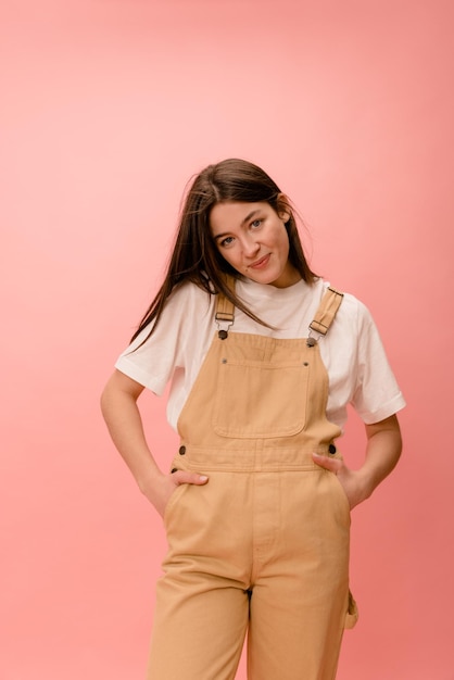 Pretty young caucasian brunette woman modestly looking at camera holding hands in pockets of overalls in studio Lifestyle leisure concept