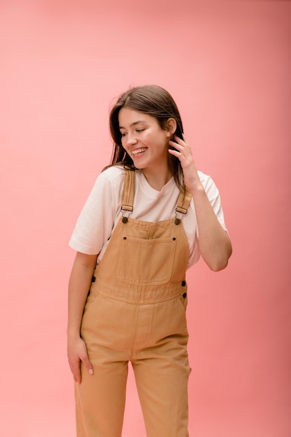 ピンクの背景に目をそらしてポーズをとるTシャツとオーバーオールのかなり若い白人ブルネットの女性人々の感情のライフスタイルとファッションのコンセプト