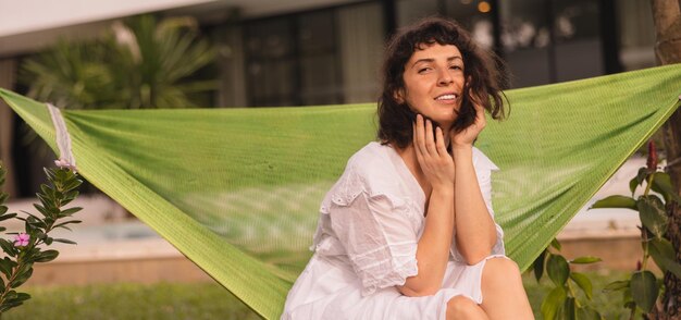 Pretty young caucasian brunette woman wear white dress spends weekend at dacha sitting in hammock summer