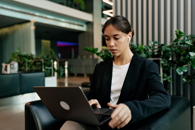 Pretty young caucasian woman is studying while sitting in modern office Appealing woman in white tshirt and dark jacket spends time doing useful activities Education concept