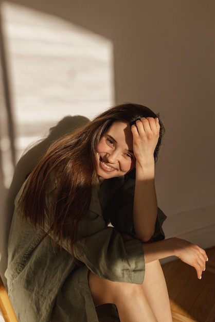 Pretty young caucasian woman looking at camera smiling broadly with teeth indoors Longhaired brunette wears long green shirt Positive emotions concept