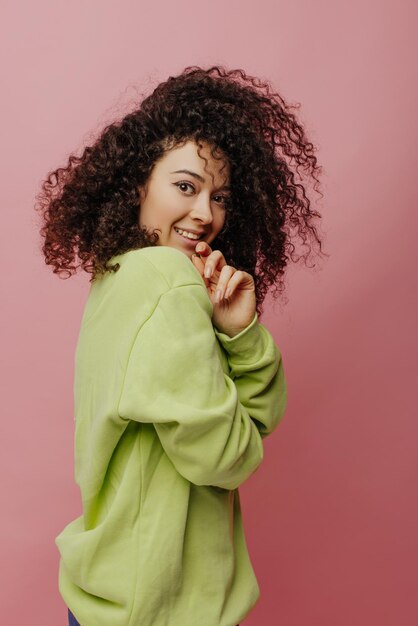 Pretty young caucasian woman with wavy hair looks piercingly into camera while standing sideways on pink background Brunette wears light green sweater Beauty and youth concept