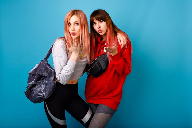 Pretty young couple of best friend girls sending air kisses, wearing fitness sports clothes and bags, healthy lifestyle, blue wall.