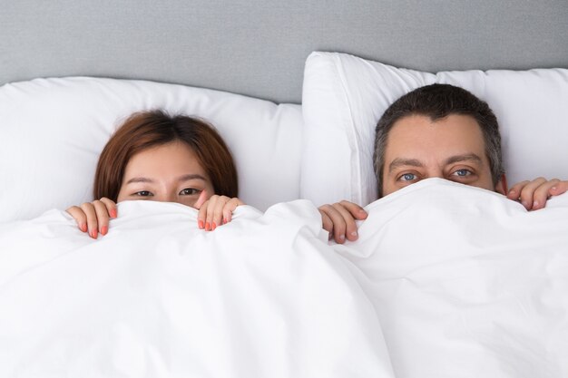 Pretty young couple hiding behind duvet