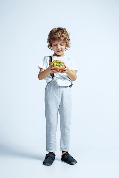 Pretty young curly boy in casual clothes on white studio wall. Eating burger