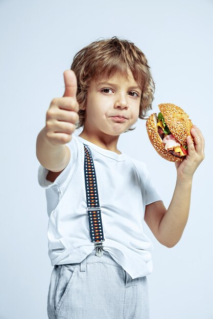 白いスタジオの壁にカジュアルな服を着たかなり若い巻き毛の少年。ハンバーガーを食べる