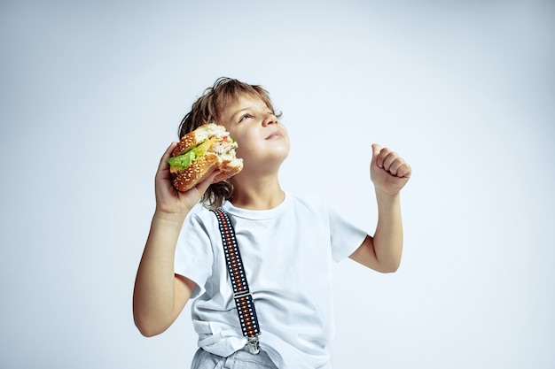 Free photo pretty young curly boy in casual clothes on white  wall. eating burger. caucasian male preschooler with bright facial emotions. childhood, expression, fun, fast food. dreamful looks up.