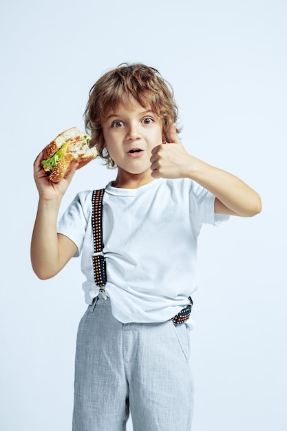 Ragazzo abbastanza giovane riccio in abbigliamento casual sulla parete bianca. mangiare hamburger. bambino in età prescolare maschio caucasico con emozioni facciali luminose. infanzia, espressione, divertimento, fast food. mostra pollice in su.