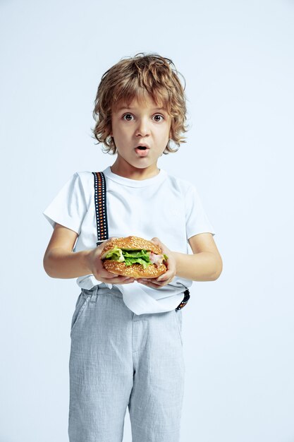 Pretty young curly boy in casual clothes on white  wall. Eating burger. Caucasian male preschooler with bright facial emotions. Childhood, expression, having fun, fast food. Astonished.
