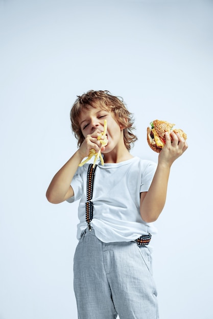 白い壁にカジュアルな服を着たかなり若い巻き毛の少年。フライドポテトと一緒にハンバーガーを食べる。明るい顔の感情を持つ白人の男性未就学児。子供時代、表現、楽しい、ファーストフード。