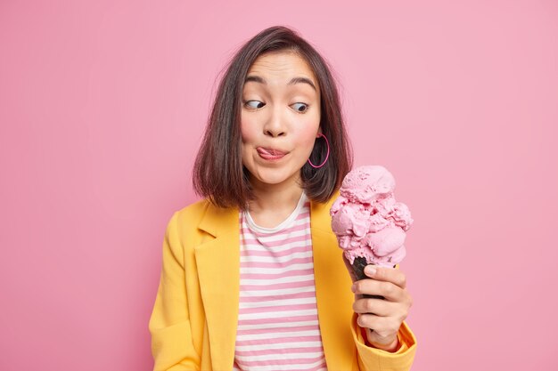 Симпатичная молодая темноволосая азиатка смотрит на аппетитное вкусное мороженое, облизывает губы языком, не может дождаться, пока съест вкусный летний десерт, одетый в модную одежду. Концепция сладкого зуба