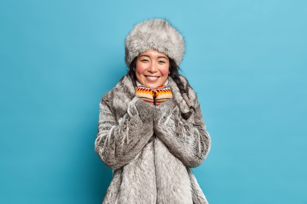 Free photo pretty young eskimo woman with rouge cheeks after spending time at frost smiles gently has two pigtails wears fur coat and hat knitted mittens isolated over blue wall