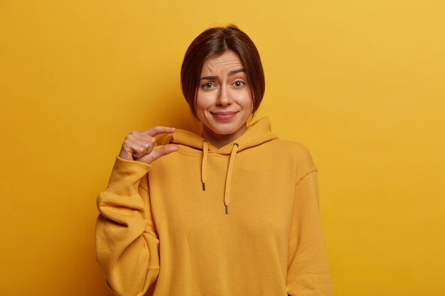 Pretty young European woman shows small size, demonstrates tiny measure, speaks about amount, dressed inn casual hoodie, shapes little object, isolated on yellow wall. Body language concept.
