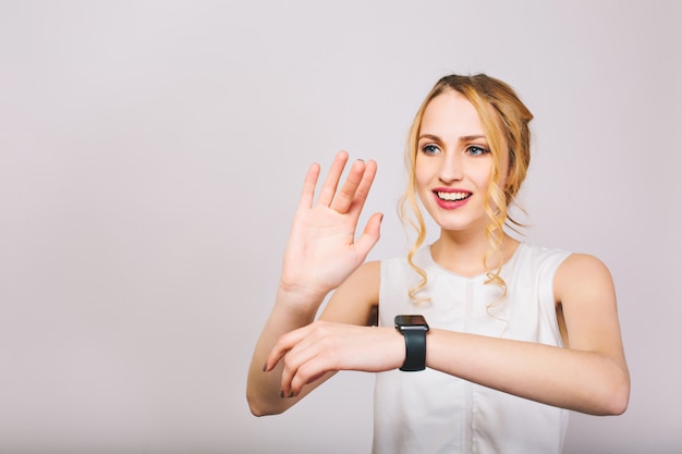 Pretty young fair-haired lady waving with her hands and posing with smile isolated. Charming girl with curly hair wearing stylish tank top laughing and showing new black wristwatch