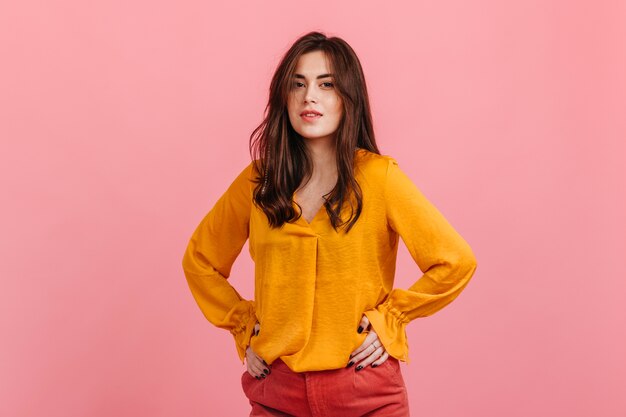 Pretty young female model with brown eyes in bright yellow shirt posing on pink wall.