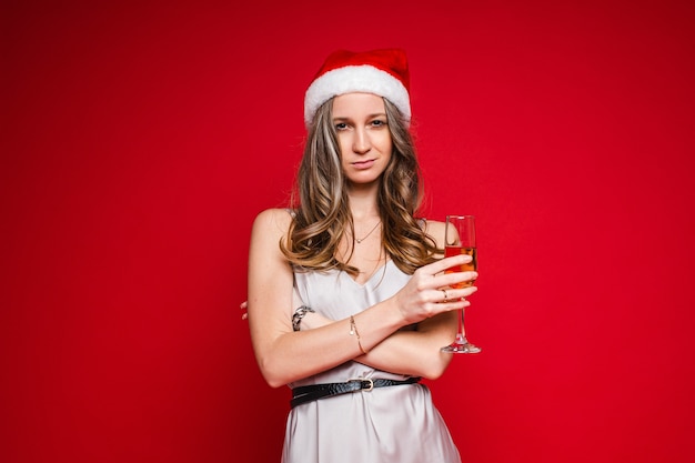 Pretty young female in santa hat and festive dress posing holding champagne glass on red background, copy space