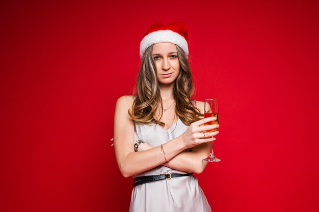 산타 모자와 빨간색 배경에 샴페인 잔을 들고 포즈를 취하는 축제 드레스에 꽤 젊은 여성, 복사 공간