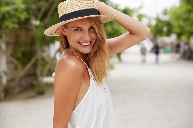 Pretty young female in summer hat and white dress, has positive expression, poses outdoor on coastline in tropical place, enjoys hot weather and sunshine. People, rest, lifestyle, season concept