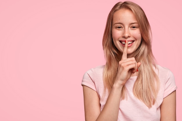 Pretty young female with cheerful expression, tells her secret to close friend, makes silence sign, stands against pink wall with copy space aside. People, privacy and secrecy concept.