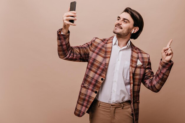 Pretty young gentleman with brunette hair and bristle wearing white shirt plaid blazer headphones holding phone video chatting and pointing at free space isolated over beige background