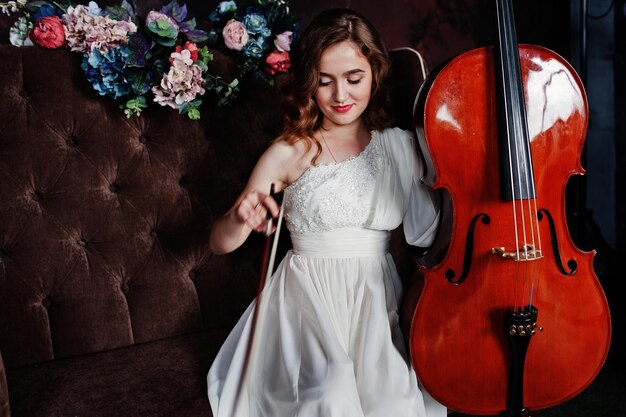 Pretty young gilrl musician in white dress with double bass sitting on brown vintage sofa