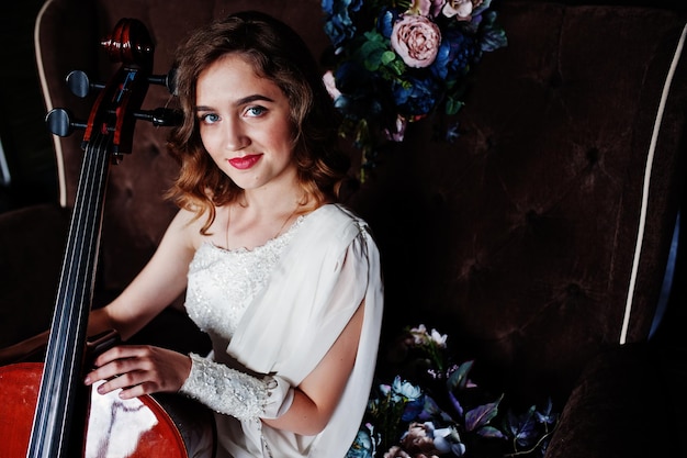 Симпатичная молодая девушка-музыкант в белом платье с контрабасом сидит на коричневом винтажном диване