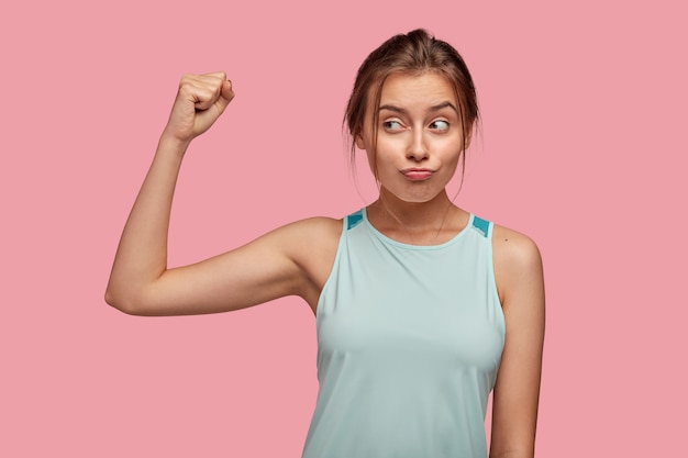 Симпатичная молодая девушка уверенная в своем теле и силе, показывает бицепс, поджимает губы, занимается спортом, носит синюю футболку, позирует у розовой стены. Здоровая спортивная женщина поднимает руку с мышцей