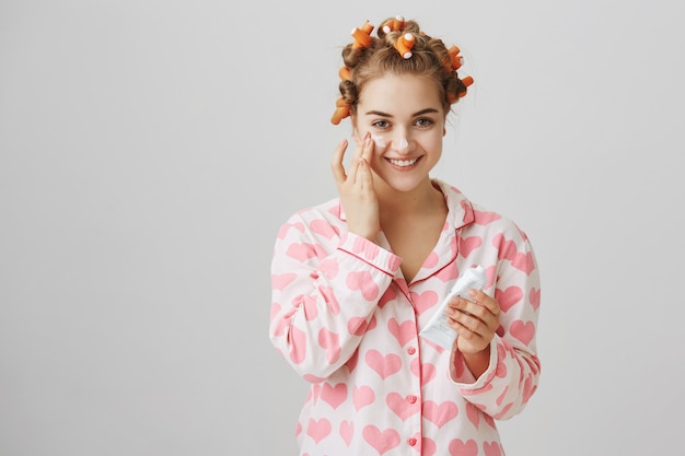Pretty young girl in hair curlers and pajama apply facial cream