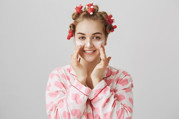 Pretty young girl in hair curlers and pajama apply facial cream