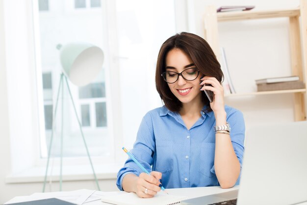 A pretty young girl is sitting at the table in office. She has blue shirt and black glasses. She is speaking on phone and writting on notebook.