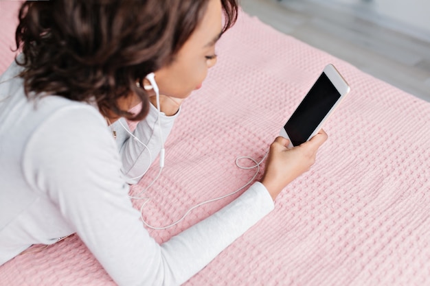 Pretty young girl lying on bed with nice pink carpet, listening to music in earphones, looking at smartphone, resting at home. Wearing light gray t-shirt with long sleeves.