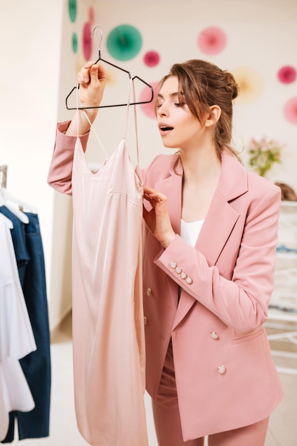 Pretty young girl in pink pantsuit have shopping in fashion boutique. Beautiful lady choose beige dress and looking with interest what price of it in clothes store