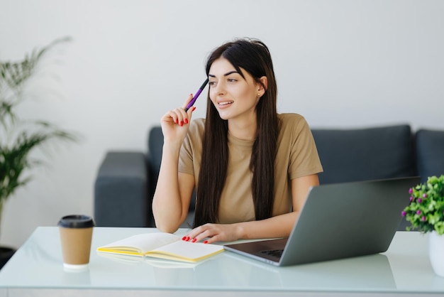 Pretty young girl sit desktop pc hold pen write notepad wear glasses shirt in home office indoors