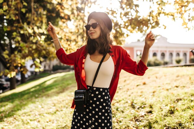 Pretty young girl with beret sunglasses white top stylish skirt and red shirt walking around early autumn city Sunny warm weather background