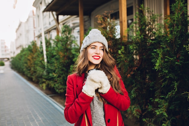 Ragazza graziosa con capelli lunghi in cappotto rosso e cappello lavorato a maglia che cammina sulla casa di legno. tiene il caffè da portare in guanti bianchi, sorridendo amichevole al lato.