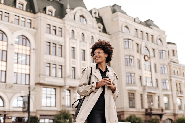 Pretty young happy brunette woman in black dress and trendy beige trench coat smiles and holds coffee cup outdoors