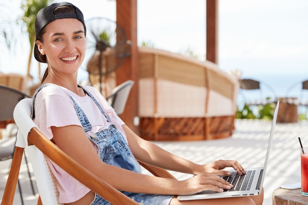 Pretty young hipster girl with pleasant smile, has positive look, dressed casualyy, rests in outdoor cafe, surfes social networks on portable laptop computer