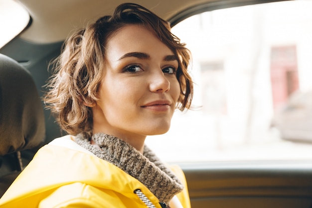 La giovane signora graziosa si è vestita in impermeabile che si siede in automobile.