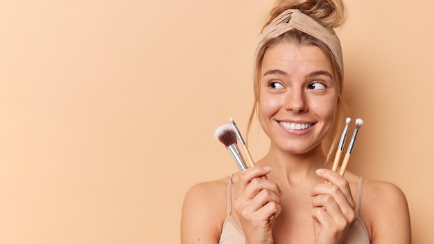 La bella giovane donna distoglie lo sguardo usa volentieri i pennelli cosmetici mette il fard applica il trucco decorativo sul viso indossa la fascia per la maglietta casual si leva in piedi con le spalle nude isolate su sfondo beige