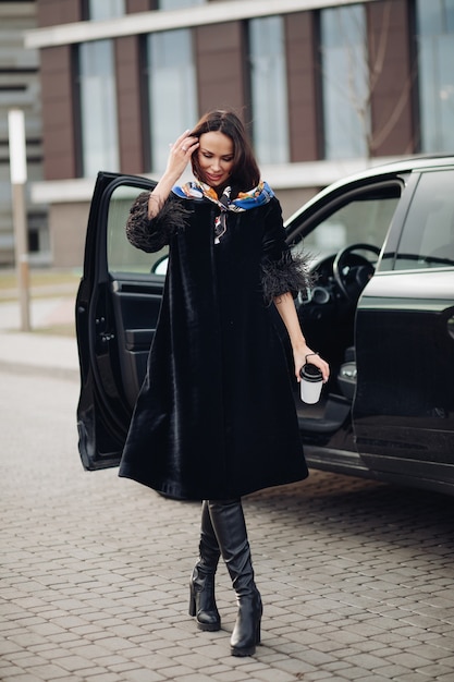 Free photo pretty young lady wearing fashionable black coat while keeping coffee and standing near car. fashion city concept