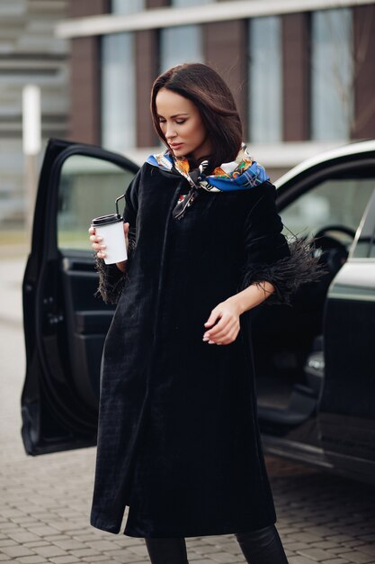 Довольно молодая дама в модном черном пальто, держа кофе и стоя возле машины. Концепция города моды