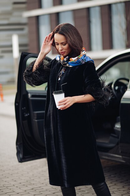 コーヒーを飲みながら車の近くに立っている間、ファッショナブルな黒いコートを着ているかなり若い女性。ファッション都市のコンセプト