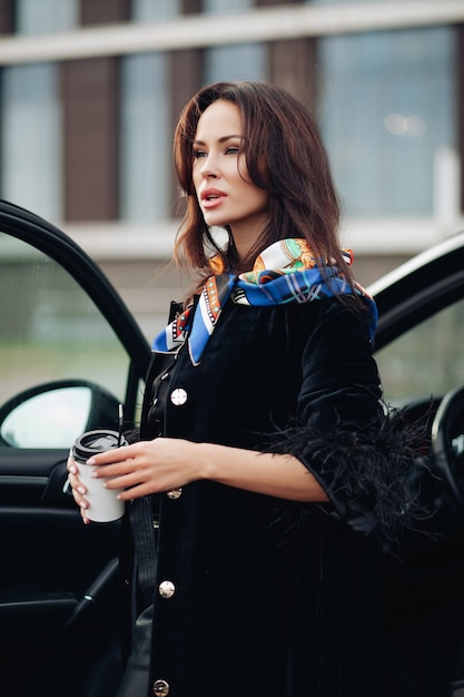 Довольно молодая дама в модном черном пальто, держа кофе и стоя возле машины. Концепция города моды