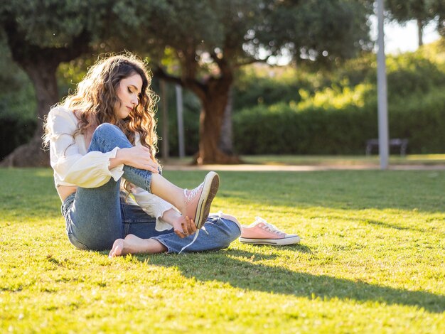 Довольно молодая длинноволосая женщина, сидящая на траве в парке, снимает кроссовки