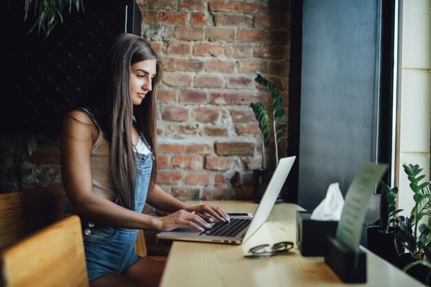 Pretty young model girl is sitting in the cafe in front of the window works on her laptop