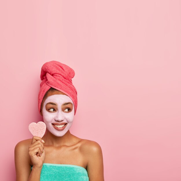 Pretty young model has well cared skin, wears facial mask for reducing dark dotes and bags under eyes, holds cosmetic sponge, wrapped bath towel on head