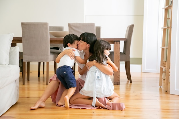 Pretty young mother sitting on floor and embracing children. Happy Caucasian mom with closed eyes kissing son and hugging daughter with love. Motherhood, weekend and parenthood concept