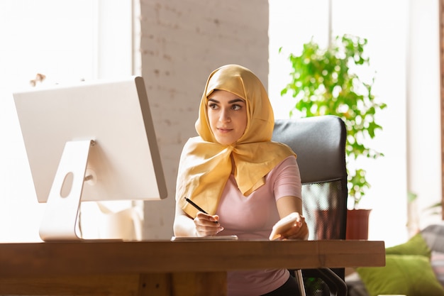 pretty young muslim woman at home during quarantine and self-insulation, using tablet for selfie or videocall, online lessons