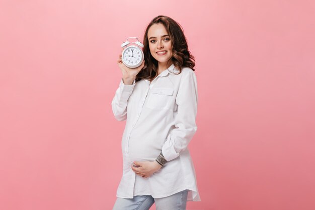 Pretty young pregnant woman in white shirt smiles widely and poses on isolated. Brunette girl holds alarm clock on pink background.