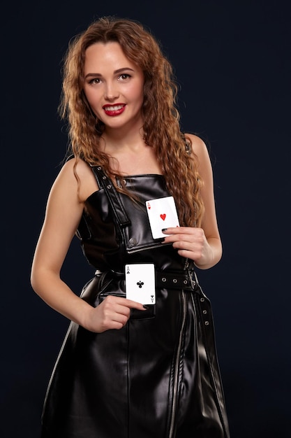 Pretty young redhead or brown-haired woman smiling, holding pair of aces wearing black latex or leather dress on black background in studio. Casino concept, gambling industry